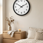 Horloge murale noire à chiffres arabes dans une chambre aux tons naturels, au-dessus d’une commode en bois avec un vase et une bougie.